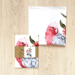 May include: White cotton handkerchiefs featuring watercolour illustrations of a pink and grey Galah cockatoo and red flowers. One handkerchief is folded, and another is packaged with a label that reads "Silken Twine, Galah Handkerchief, 34cm x 34cm, 100% cotton."