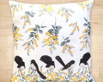 May include: White throw pillow with a floral pattern of yellow wattle flowers and green leaves. Five black and white birds are perched on the branches.