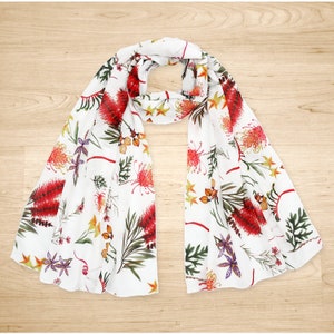 May include: White silk scarf with a floral print featuring red, yellow, and green flowers. The scarf is folded in half and laid flat on a wooden surface.