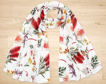 Puede incluir: Bufanda de seda blanca con estampado floral con flores rojas, amarillas y verdes. La bufanda está doblada por la mitad y colocada sobre una superficie de madera.
