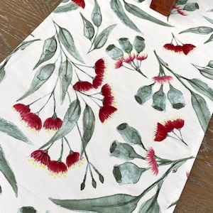 May include: A white table runner with a repeating pattern of red gum blossoms and green eucalyptus leaves. The floral design is painted in a watercolor style.