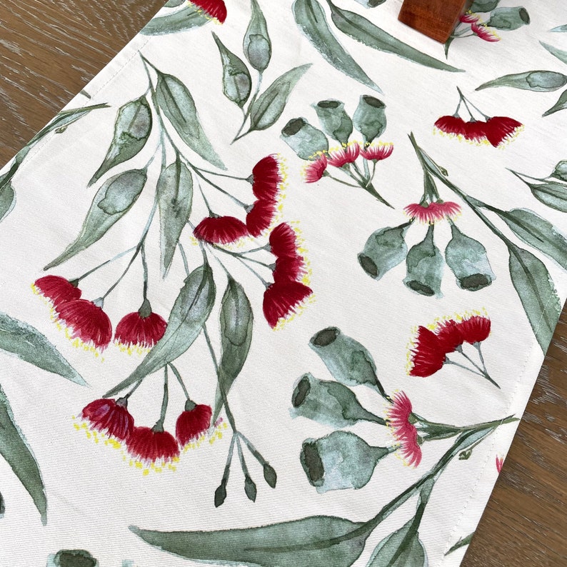 May include: A white table runner with a repeating pattern of red gum blossoms and green eucalyptus leaves. The floral design is painted in a watercolor style.
