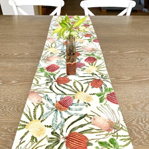 May include: A white table runner with a floral pattern of red, pink, and yellow flowers. The runner is on a wooden table with two white chairs in the background.