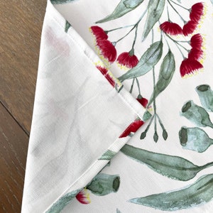 May include: A white table runner with a watercolor design of red gum blossoms and green eucalyptus leaves.