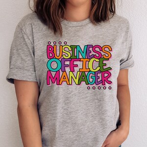 以下が含まれることがあります： カラフルな文字で「BUSINESS OFFICE MANAGER」と書かれたグレーのTシャツです。