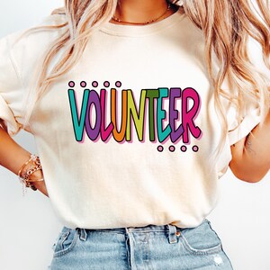 May include: A cream-colored short-sleeve t-shirt featuring the word VOLUNTEER printed in a multi-colored, bold, rounded font. The letters are colored in teal, purple, orange, yellow, green, and pink, with small circles above and below the text.