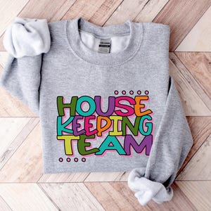 May include: Grey sweatshirt with the text "HOUSEKEEPING TEAM" in colourful, outlined letters.