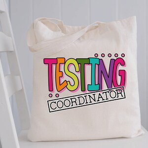 Op de afbeelding: Cr&egrave;mekleurige draagtas met de woorden "TESTING COORDINATOR" in kleurrijke, vette letters. De tekst is zwart omlijnd en heeft een verscheidenheid aan heldere kleuren, waaronder roze, oranje, groen en blauw. De tas staat op een witte stoel.