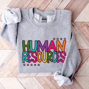 May include: A heather gray sweatshirt with the words "HUMAN RESOURCES" in a colorful, block letter design. The text is outlined in black and filled with shades of green, purple, orange, and pink. The sweatshirt has a crew neck and long sleeves.