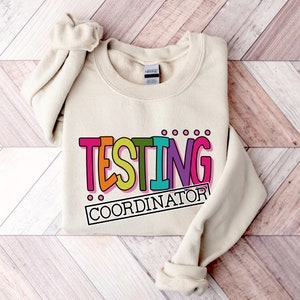 Op de afbeelding: Een cr&egrave;mekleurige sweatshirt met de tekst "TESTING COORDINATOR" in een kleurrijk, speels lettertype. De tekst is zwart omlijnd en heeft roze, oranje, turquoise en paarse tinten. De sweatshirt ligt op een houten oppervlak.