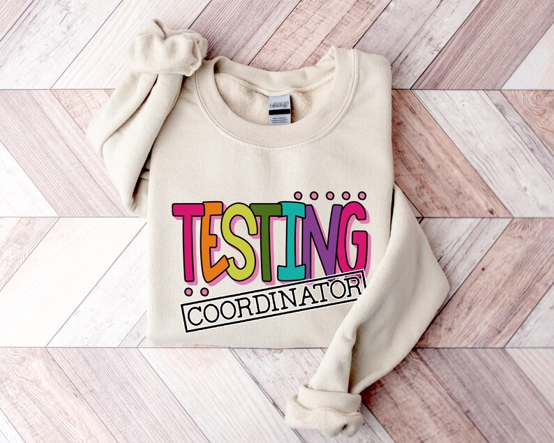 Op de afbeelding: Een cr&egrave;mekleurige sweatshirt met de tekst "TESTING COORDINATOR" in een kleurrijk, speels lettertype. De tekst is zwart omlijnd en heeft roze, oranje, turquoise en paarse tinten. De sweatshirt ligt op een houten oppervlak.