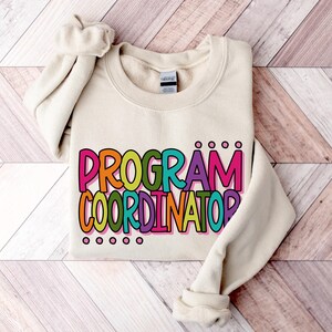 May include: Cream-colored sweatshirt with the words "PROGRAM COORDINATOR" in large, colorful letters. The text is outlined in black and filled with shades of orange, green, purple, and pink. The sweatshirt is folded on a light wood-grain surface.