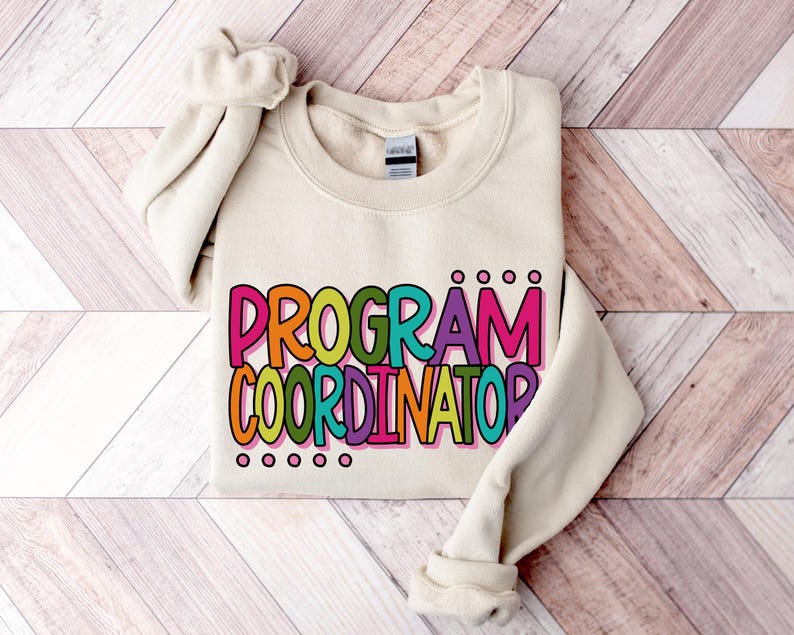 May include: Cream-colored sweatshirt with the words "PROGRAM COORDINATOR" in large, colorful letters. The text is outlined in black and filled with shades of orange, green, purple, and pink. The sweatshirt is folded on a light wood-grain surface.