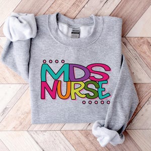 May include: A heather gray crewneck sweatshirt with the text "MDS NURSE" in colorful, outlined letters. The design includes teal, pink, orange, green, and purple. The sleeves are folded up. The sweatshirt is on a wooden surface.
