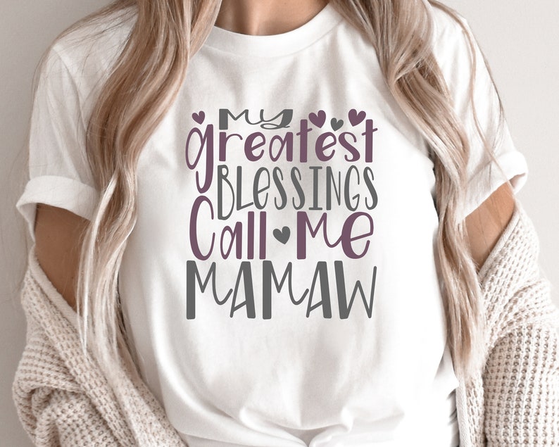 Peut inclure: T-shirt blanc avec le texte "My greatest blessings call me Mamaw" en lettres grises et violettes avec des c&oelig;urs.