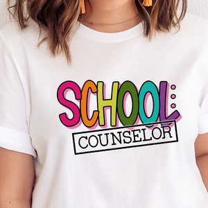 以下が含まれることがあります： カラフルな輪郭のある文字で「SCHOOL COUNSELOR」と書かれた白いTシャツ。 「SCHOOL」という単語は虹色の色で、「COUNSELOR」という単語は黒い長方形の中にあります。