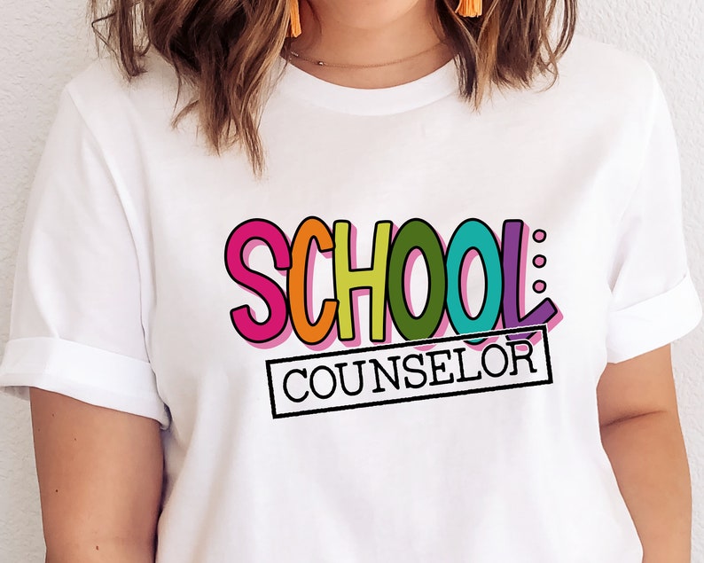 以下が含まれることがあります： カラフルな輪郭のある文字で「SCHOOL COUNSELOR」と書かれた白いTシャツ。 「SCHOOL」という単語は虹色の色で、「COUNSELOR」という単語は黒い長方形の中にあります。