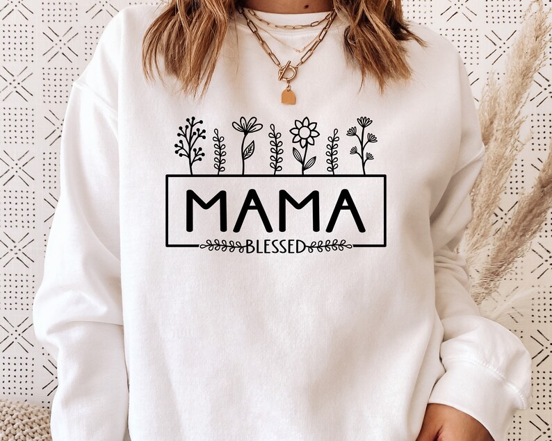 May include: White sweatshirt with black text that reads "MAMA" in a rectangular box with a floral design above and the word "BLESSED" below.