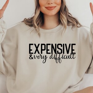 Może przedstawiać: Beżowa bluza z nadrukiem "EXPENSIVE & very difficult" w kolorze czarnym.