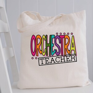 May include: A white canvas tote bag with the words "ORCHESTRA TEACHER" in colorful letters. The word "ORCHESTRA" is in a rainbow of colors and the word "TEACHER" is in a black rectangle.