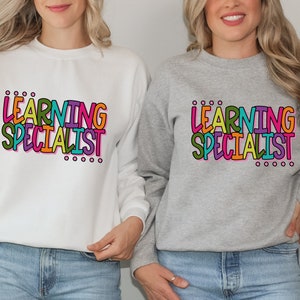 May include: Two women wearing grey and white sweatshirts with the text "LEARNING SPECIALIST" in colorful letters.