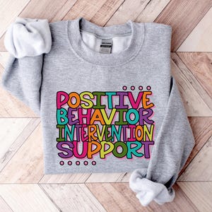 Könnte beinhalten: Ein meliertes graues Sweatshirt mit dem Text "POSITIVE BEHAVIOR INTERVENTION SUPPORT" in einem bunten Blockbuchstaben-Design. Der Text ist schwarz umrandet und hat rosa, blaue, grüne, gelbe und violette Farben. Das Sweatshirt liegt auf einer Holzoberfläche.