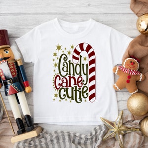 May include: White t-shirt with a red and white candy cane and green text that says "Candy Cane Cutie".
