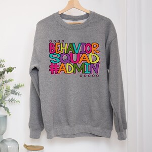 May include: A heather gray sweatshirt with the words "BEHAVIOR SQUAD #ADMIN" in a colorful, blocky font. The text is in shades of orange, pink, green, yellow, and purple. The sweatshirt is hanging on a wooden hanger.