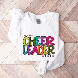 May include: White sweatshirt with the words "CHEER LEADER" in colorful, outlined letters. The letters are in different colors, including pink, green, yellow, and orange.