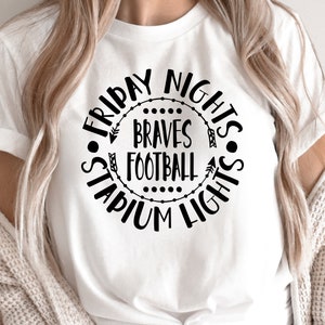 Puede incluir: Camiseta blanca con un diseño circular negro que dice "Friday Nights Braves Football Stadium Lights". El diseño incluye flechas y puntos. La camiseta está hecha de un material suave.