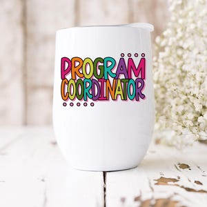 May include: A white insulated wine tumbler with a lid. The tumbler features the words "PROGRAM COORDINATOR" in colorful, bubble-style letters. The design includes a variety of colors, including pink, orange, green, and purple. The tumbler is on a white wooden surface.