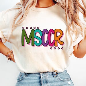 May include: A cream-colored short-sleeve t-shirt featuring the text MSCCR in a bold, multi-colored font. Each letter is outlined in pink, with the M in green, S in teal, C in purple, C in pink, and R in orange. Small circles are above and below.
