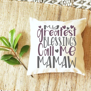 Peut inclure: Un coussin blanc avec le texte "My greatest blessings call me Mamaw" en lettres grises et violettes avec de petits c&oelig;urs.