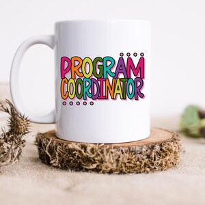 May include: A white ceramic mug with the words "PROGRAM COORDINATOR" in colorful bubble letters. The mug sits on a small, round wooden coaster. The text is in pink, orange, green, blue, and purple.