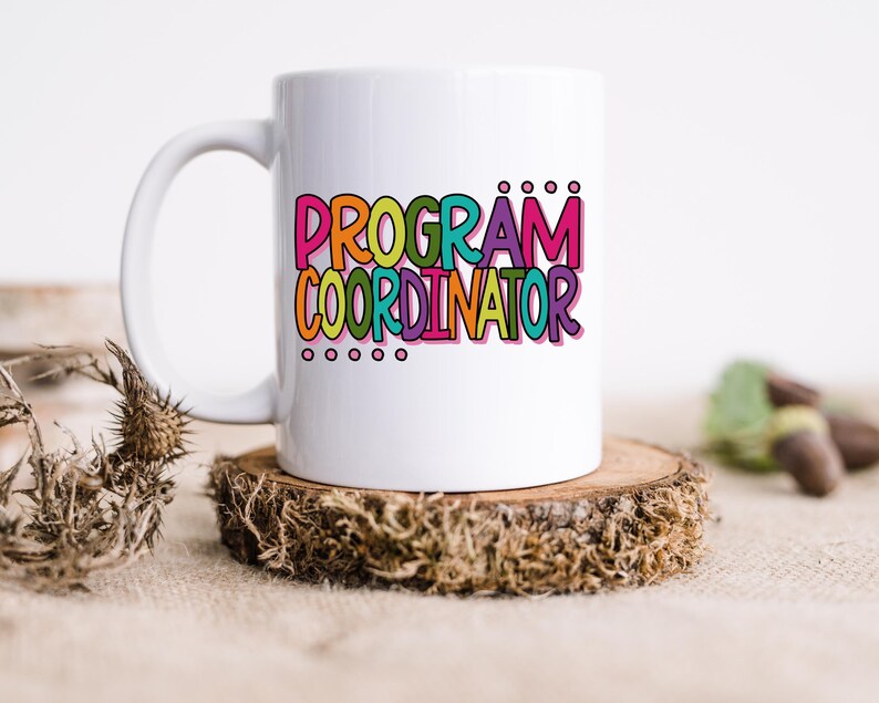 May include: A white ceramic mug with the words "PROGRAM COORDINATOR" in colorful bubble letters. The mug sits on a small, round wooden coaster. The text is in pink, orange, green, blue, and purple.