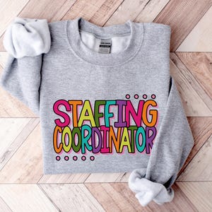 May include: A heather gray sweatshirt with the words "STAFFING COORDINATOR" in a colorful, bubble letter font. The text is outlined in black and has a playful, eye-catching design. The sweatshirt is made of a soft material.