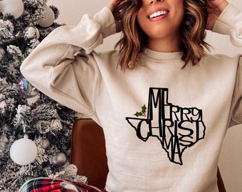 Puede incluir: Una sudadera de color crema con un contorno negro del estado de Texas. Las palabras "Merry Christmas" están escritas dentro del contorno del estado.