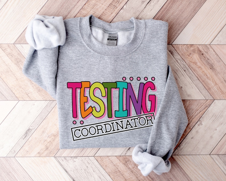 Op de afbeelding: Een grijze sweatshirt met de tekst "TESTING COORDINATOR" in kleurrijke, dikgedrukte letters. De tekst is zwart omlijnd en heeft een speelse, cartoonachtige stijl. De sweatshirt ligt op een lichtbruin houten oppervlak.