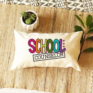 以下が含まれることがあります： カラフルな文字で「SCHOOL COUNSELOR」と書かれた白い枕。枕は、緑の植物が入った鉢のある織り込まれた敷物の上に置かれています。