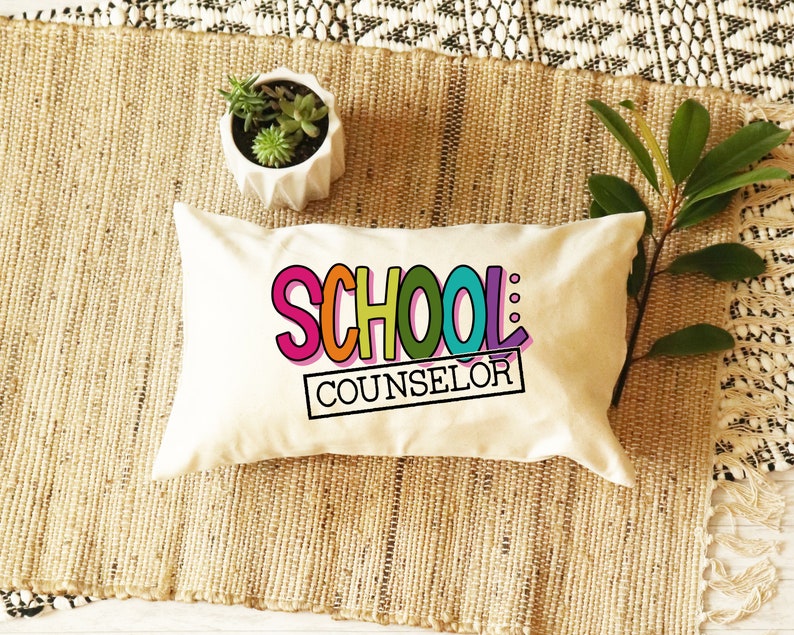 以下が含まれることがあります： カラフルな文字で「SCHOOL COUNSELOR」と書かれた白い枕。枕は、緑の植物が入った鉢のある織り込まれた敷物の上に置かれています。