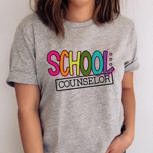 以下が含まれることがあります： カラフルな文字で「SCHOOL: COUNSELOR」と書かれたグレーのTシャツ。 「SCHOOL」という単語は虹色の色で、「COUNSELOR」という単語は黒い長方形の中にあります。