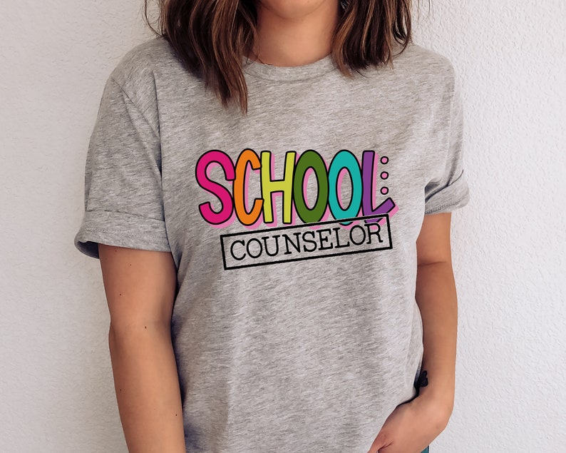 以下が含まれることがあります： カラフルな文字で「SCHOOL: COUNSELOR」と書かれたグレーのTシャツ。 「SCHOOL」という単語は虹色の色で、「COUNSELOR」という単語は黒い長方形の中にあります。