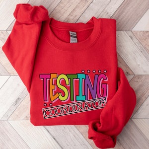 Op de afbeelding: Een felrood sweatshirt met de tekst "TESTING COORDINATOR" in kleurrijke letters. De tekst is zwart omlijnd en heeft een retro, speels ontwerp. Het sweatshirt is gemaakt van een zacht materiaal.