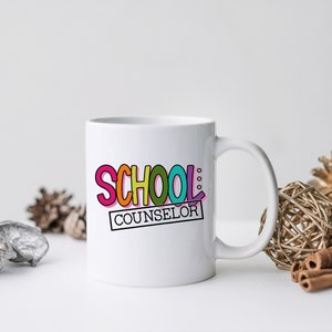 以下が含まれることがあります： 虹色のフォントで「SCHOOL COUNSELOR」と書かれたカラフルなグラフィックが施された白いセラミックマグカップです。