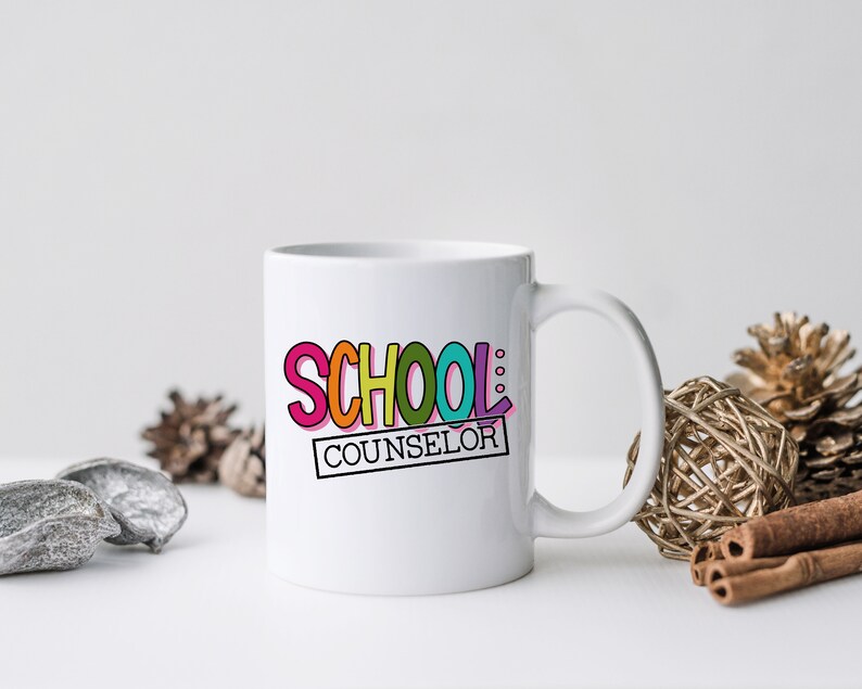 以下が含まれることがあります： 虹色のフォントで「SCHOOL COUNSELOR」と書かれたカラフルなグラフィックが施された白いセラミックマグカップです。