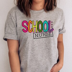 May include: A gray t-shirt with the text "SCHOOL: NURSE" in colorful letters. The letters are outlined in black and have a playful, cartoon-like style.