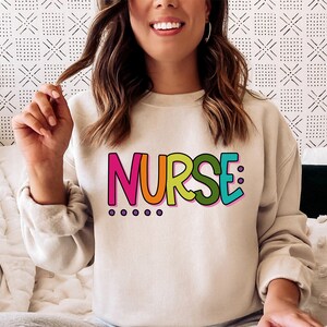 Peut inclure: Un sweat-shirt blanc avec le mot "NURSE" imprim&eacute; en lettres color&eacute;es. Les lettres sont soulign&eacute;es de rose et ont un sch&eacute;ma de couleurs arc-en-ciel.
