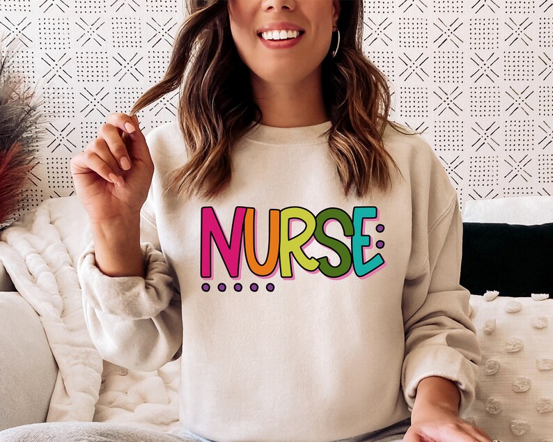 Peut inclure: Un sweat-shirt blanc avec le mot "NURSE" imprim&eacute; en lettres color&eacute;es. Les lettres sont soulign&eacute;es de rose et ont un sch&eacute;ma de couleurs arc-en-ciel.