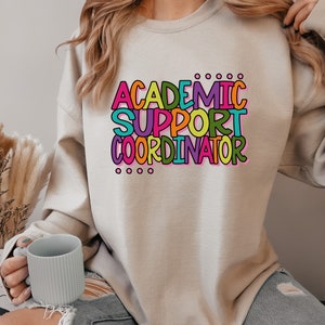 May include: A light beige sweatshirt with the text "ACADEMIC SUPPORT COORDINATOR" in colorful letters. The letters are in different colors, including pink, yellow, green, blue, and orange.