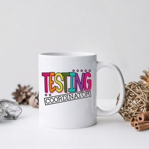 Op de afbeelding: Witte keramische mok met de tekst "TESTING COORDINATOR" in kleurrijke, dikgedrukte letters. De mok heeft een gebogen handvat en staat tegen een neutrale achtergrond met decoratieve elementen.